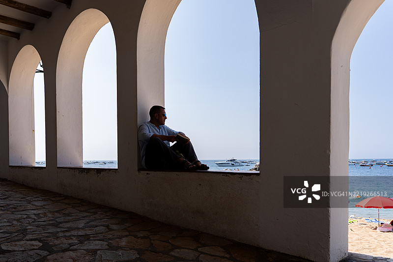 成熟男性在白色拱形窗旁悠闲地阅读书籍，凝视着海景。图片素材
