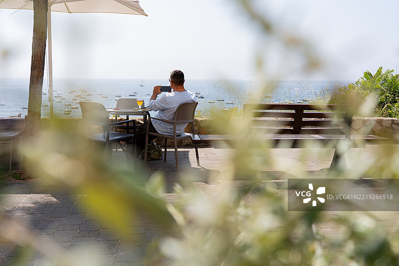 商人在餐厅享用饮品的同时，用智能手机拍摄海景。图片素材