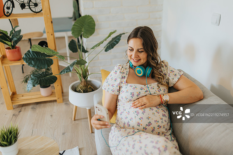 快乐的孕妇在舒适的家中沙发上放松时使用智能手机图片素材