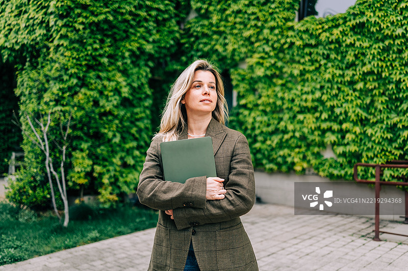 微笑的女商人手持笔记本电脑站在绿色建筑前图片素材
