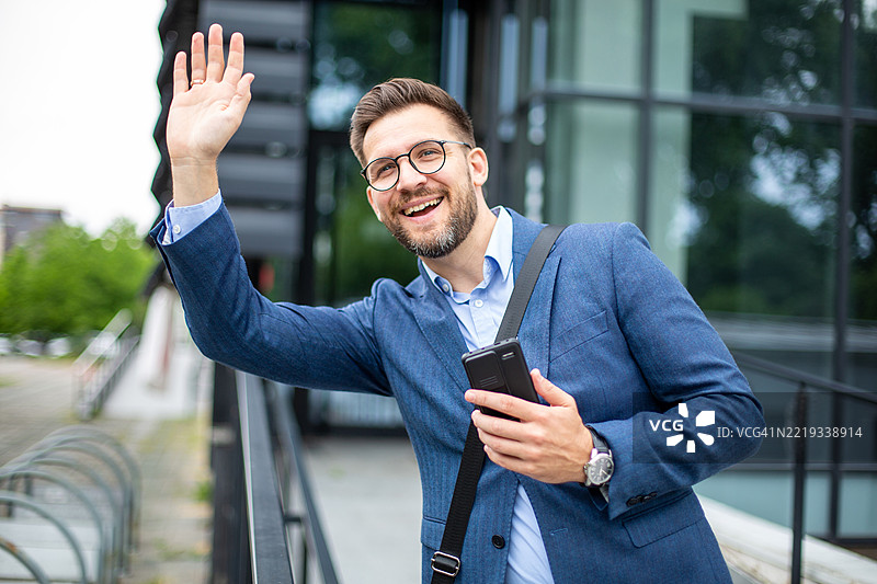 微笑的商人在城市中挥手并拿着智能手机图片素材