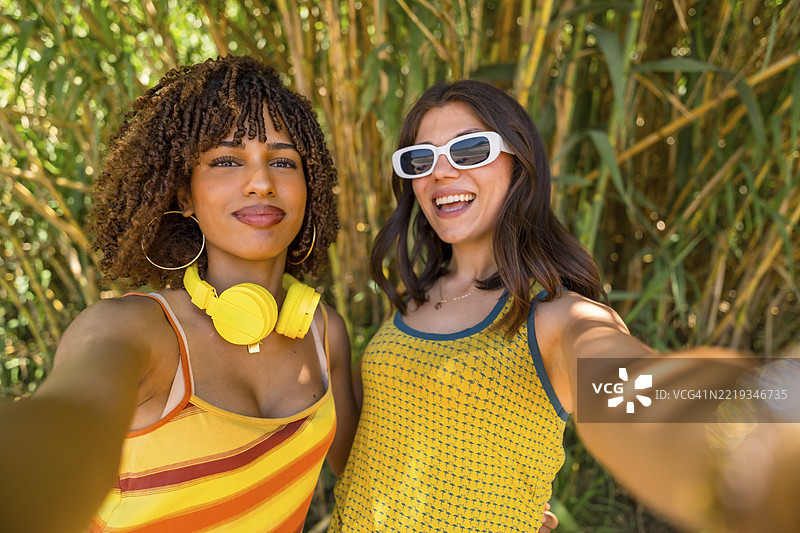 两位穿着色彩鲜艳衣服、戴着耳机的年轻女性在竹林前自拍，面带笑容。图片素材