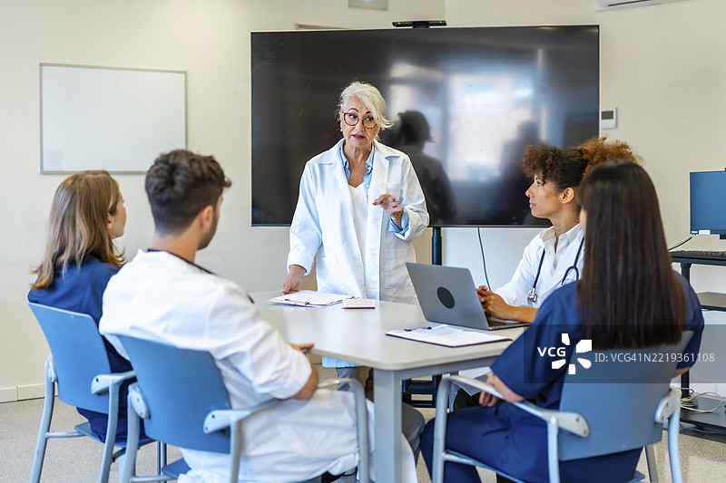 经验丰富的医生在医学培训中向同事解释一种新治疗方法，使用笔记本电脑和显示器。图片素材