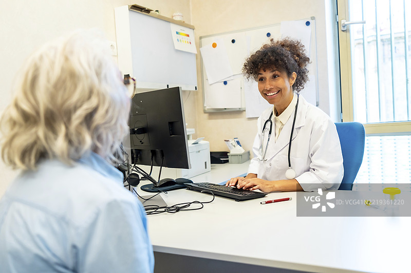 微笑的女医生穿着白大褂和听诊器，一边在电脑键盘上打字，一边倾听住院的老年女性患者。图片素材