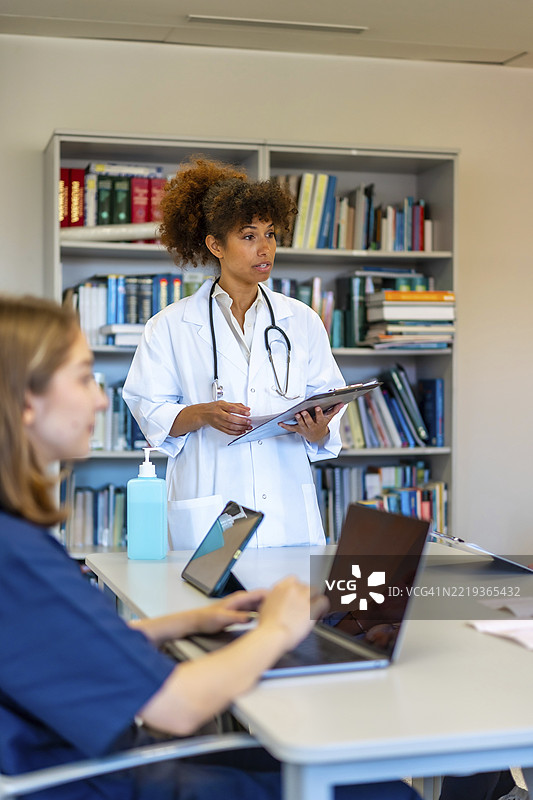 医生和护士在医疗简报中使用笔记本电脑和平板电脑，讨论病人病历和报告，一位持有夹板的高级医生参与讨论。图片素材