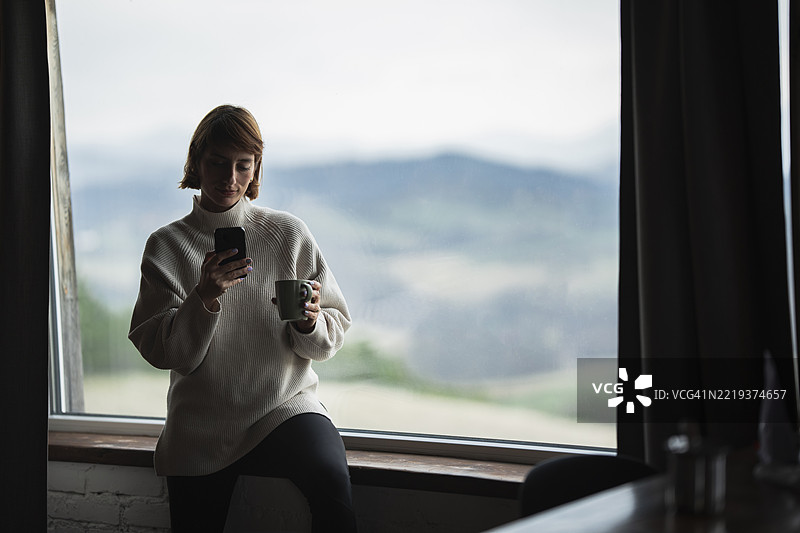 年轻女性在窗边享受咖啡，使用智能手机，窗外风景如画图片素材