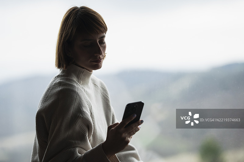 年轻女性在窗边使用智能手机，俯瞰山景。图片素材