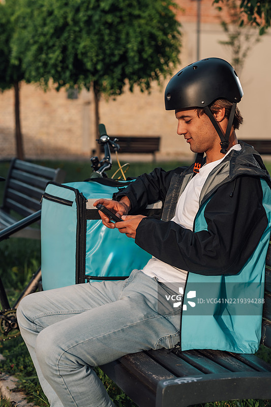 送货员坐在长椅上，查看他的智能手机，身旁有自行车和背包。图片素材