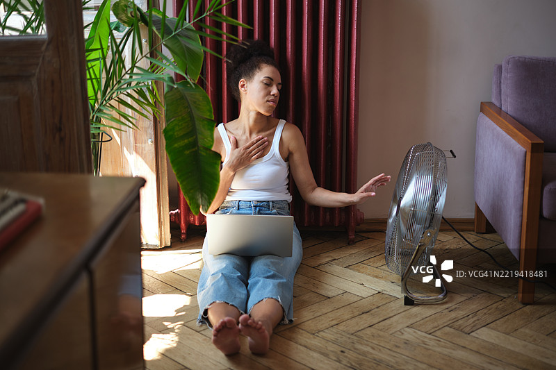 在热浪中居家工作的女性自由职业者，坐在电风扇旁边用笔记本电脑工作，感到不适。图片素材