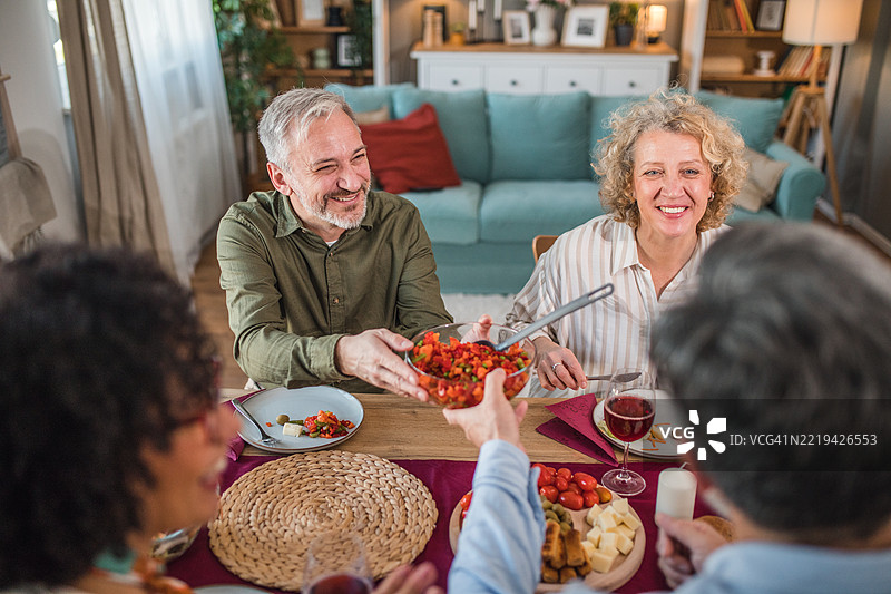 聚会的男主人将一碗沙拉递给坐在餐桌另一侧的朋友们图片素材