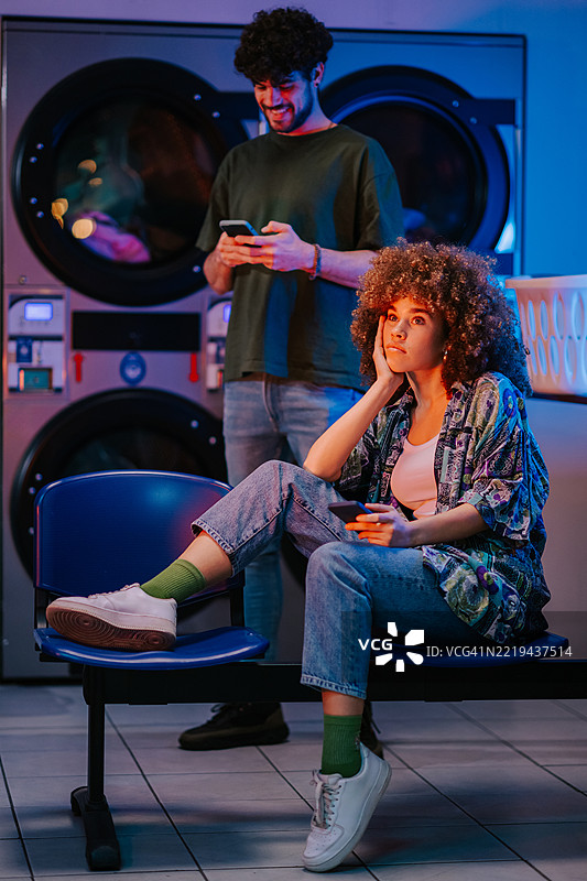 年轻情侣在自助洗衣店用智能手机等待洗衣服图片素材