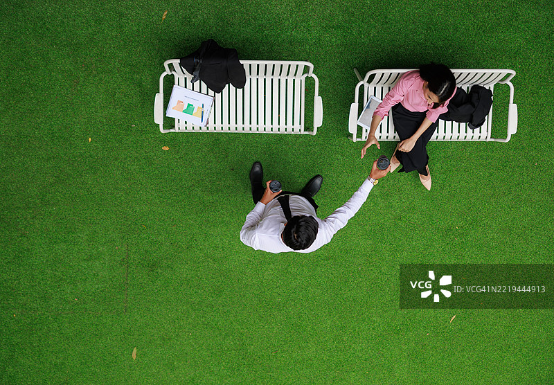 从上方俯视两位商务专业人士在公园长椅上享受咖啡休息，周围绿意盎然，工作期间进行非正式交谈，营造出促进健康与联系的城市公园环境。图片素材