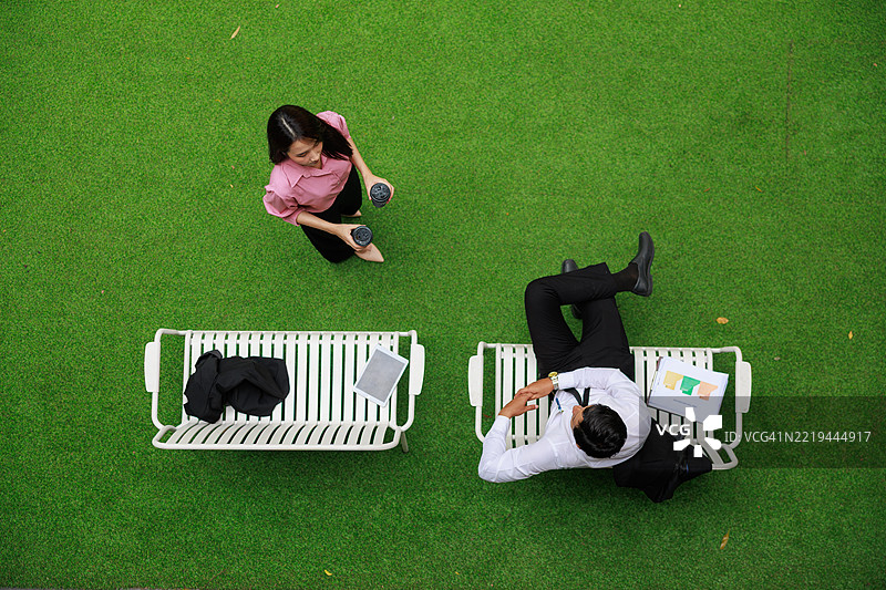 从上方俯视两位商务专业人士在公园长椅上休息，周围绿意盎然，享受着工作时间的非正式对话，营造出促进健康与联系的城市公园环境。图片素材