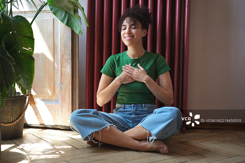 年轻的非裔美国女性坐在莲花坐姿中，双眼闭合，周围环绕着绿色植物和温暖的阳光，象征着和谐、感恩与健康，双手放在胸前微笑。代表Z世代与心理健康。图片素材