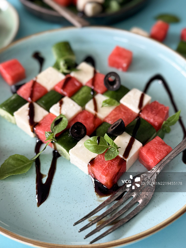 特写镜头：蓝色盘子里的西瓜希腊沙拉，交替放置着西瓜块、羊奶酪和黄瓜，点缀着黑橄榄片和薄荷叶，淋上香醋，金属叉子，前景重点突出，健康饮食。图片素材
