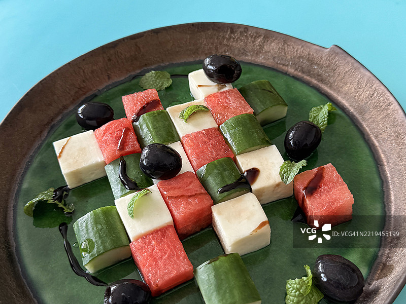 全景图像展示了一盘绿色和棕色的西瓜希腊沙拉，沙拉中交替放置着西瓜块、羊奶酪和黄瓜，点缀着黑橄榄片和薄荷叶，淋上了香醋，从高处俯视，健康饮食。图片素材