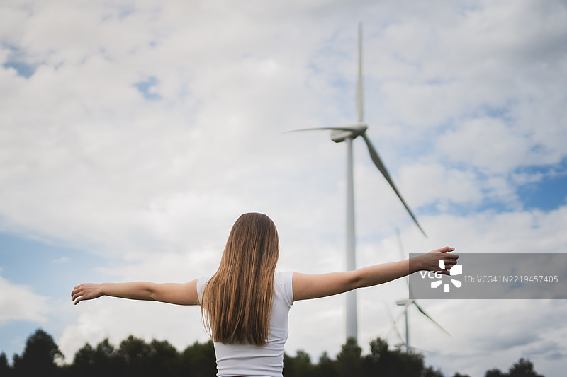 年轻女性张开双臂，仰望田野中的风力发电机图片素材