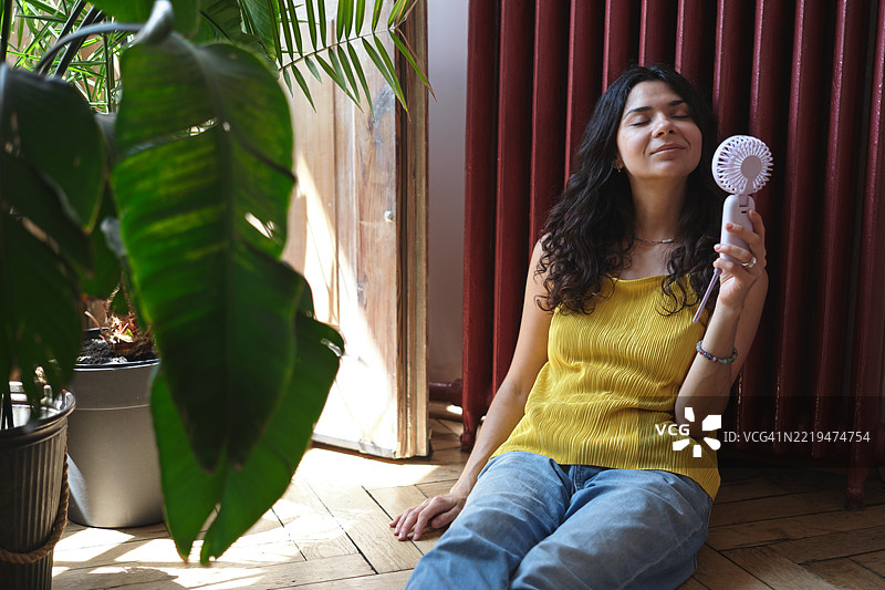 夏季炎热，生活在没有空调的环境中。年轻女性在家窗边坐着，使用便携式电动迷你风扇对着脸，享受炎热天气中的清新空气，降温。图片素材