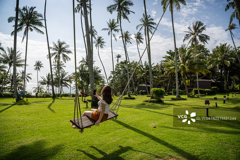 在热带棕榈树下的秋千上放松的女人，手中拿着手机，夕阳西下。图片素材
