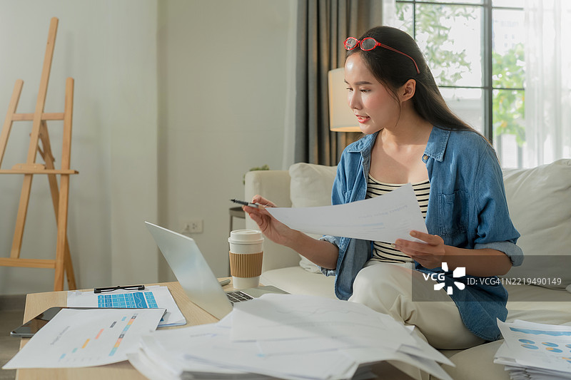 一位穿着休闲服的快乐亚洲女性在家中远程工作，舒适地坐在公寓的沙发上，使用笔记本电脑。她参加在线商务会议，同时利用人工智能助手提高工作效率，享受智能工作的乐趣。图片素材