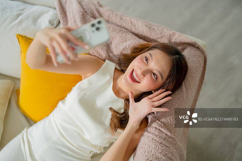 一位快乐、聪明的亚洲女性戴着眼镜，穿着休闲服，舒适地坐在沙发上使用智能手机。在人工智能的帮助下，她查看旅行信息并安排周末旅行计划，享受现代数字生活方式。图片素材