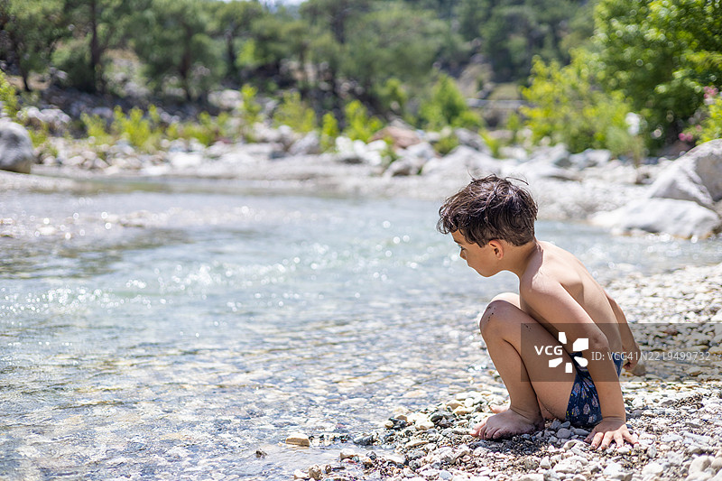 一个年轻男孩蹲在河边，享受着大自然的宁静时刻，观察着水流。图片素材