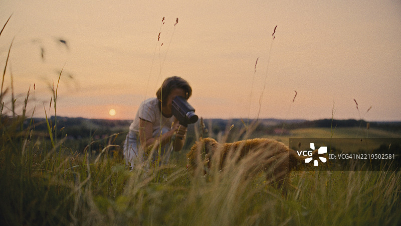 在乡村草地上，日落时分，一位女性用复古摄像机拍摄她的宠物狗。图片素材