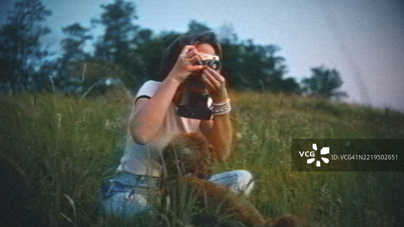 在夏季草地上，拿着老式相机的女人与宠物狗一起。模拟胶卷，VHS风格。图片素材