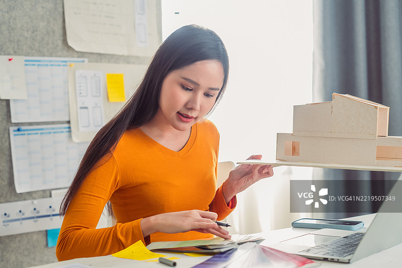 一位专注的亚洲女性建筑师在家中办公室微笑着工作，使用智能手机查看蓝图并协调新的房屋设计项目。她被绘图工具和现代装饰环绕，平衡着创造力与科技。图片素材