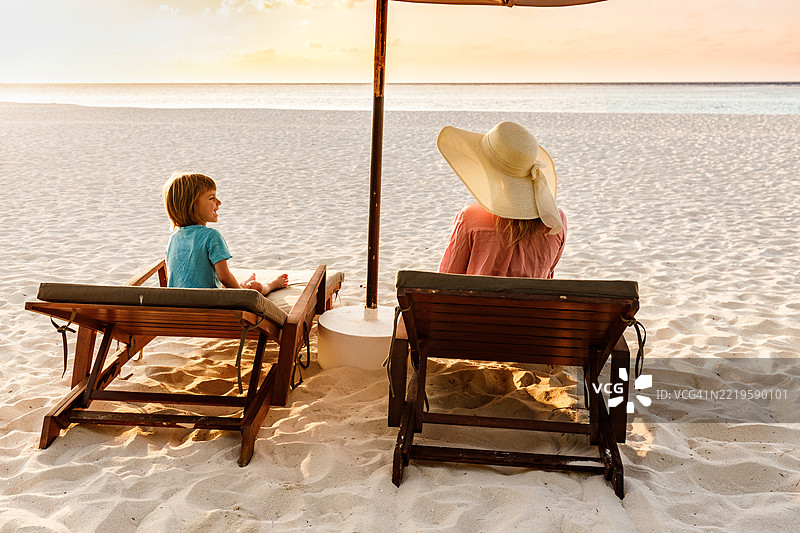 快乐的男孩和他的母亲在海滩的躺椅上放松的背影。图片素材