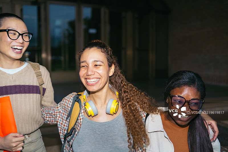 三位不同背景的大学朋友并肩走在校园里，微笑着交谈，洋溢着共同的快乐和友谊。图片素材
