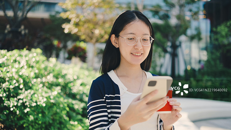 亚洲Z世代年轻女性在现代绿色公园中一边喝咖啡一边使用智能手机，背景是充满活力的城市。工作与生活的平衡、休闲和城市生活。图片素材