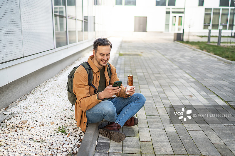 微笑的学生在户外使用智能手机并享用糕点图片素材