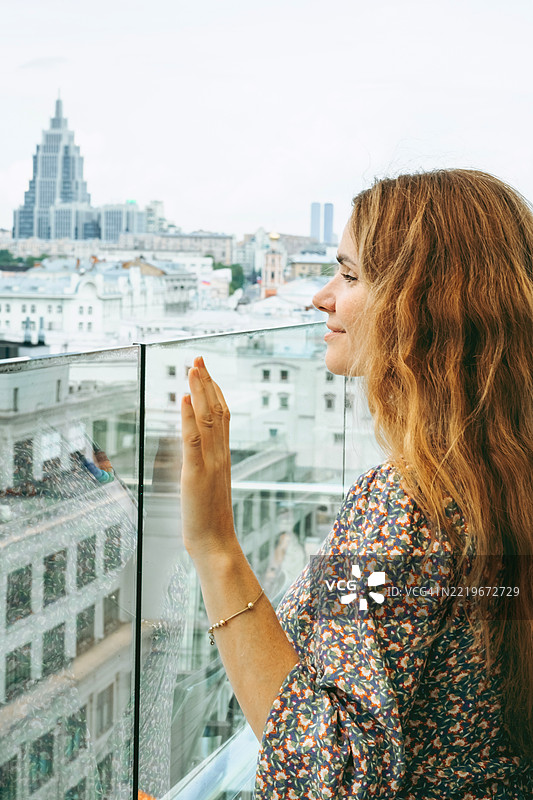 年轻的白人女性，长长的波浪形金发，透过玻璃屏障凝视城市天际线。城市背景中有高楼大厦和柔和的日光。图片素材