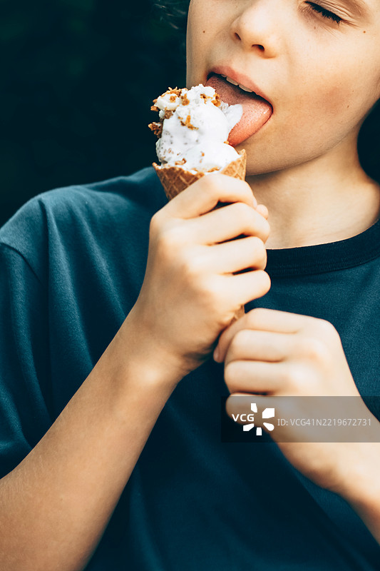 小男孩舔着装有碎屑的白色冰淇淋华夫筒。他穿着深蓝色的衬衫。背景模糊且昏暗，拍摄于自然光下的户外。图片素材