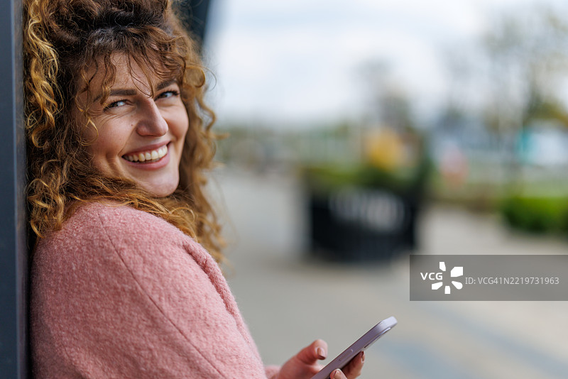 阳光明媚的城市中，微笑的女性在等待朋友时上网冲浪图片素材