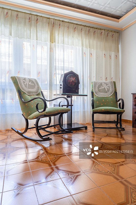 复古风格的桌面收音机放置在两把摇椅之间，摇椅上铺有钩针花边，整个空间位于一个复古的客厅中，自然光透过薄纱窗帘洒入，营造出温馨而怀旧的氛围。图片素材