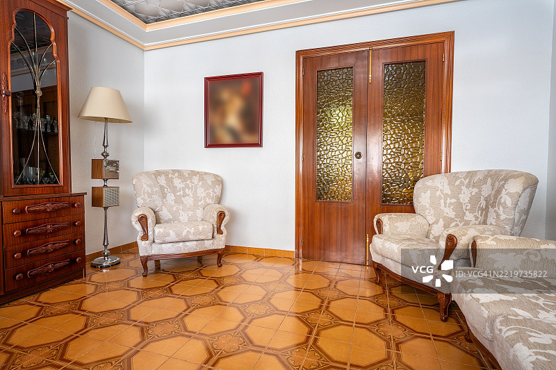宽阔视角的复古客厅，展示了两把优雅的扶手椅和一张舒适的沙发，一个木质橱柜，一盏落地灯，以及一个图案瓷砖地板，营造出1980年代的复古氛围。图片素材