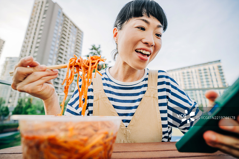 一位亚洲女性在街头咖啡馆吃面条时使用手机。图片素材