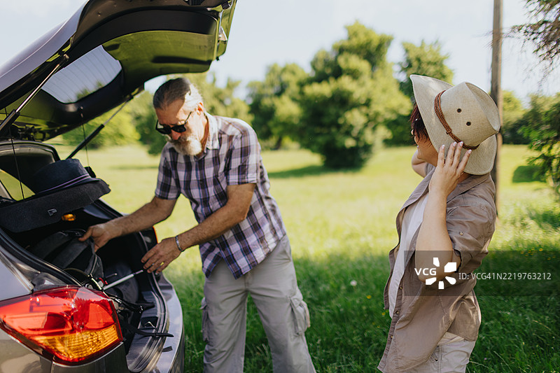 成熟夫妇站在汽车后备厢旁，准备在春季大自然中徒步旅行，戴上帽子。图片素材