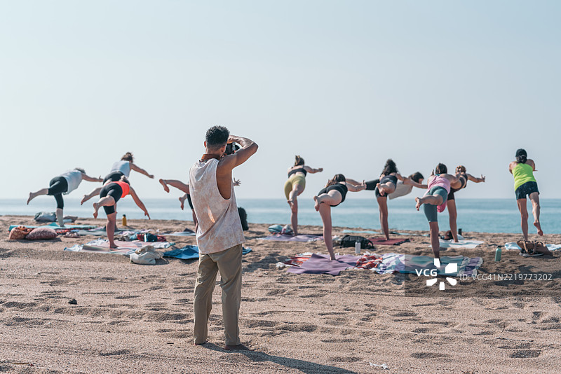 瑜伽教练在海滩上拍摄他的学生练习战士三式。图片素材