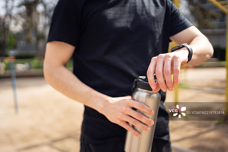 年轻男子在户外调整智能手表，手中拿着水瓶图片素材