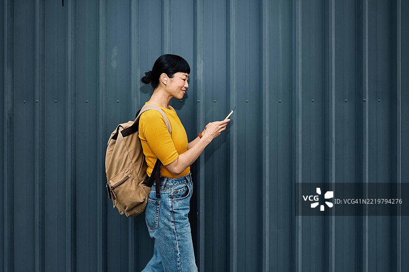 侧面视角的微笑跨种族女性在街上手持手机行走。图片素材