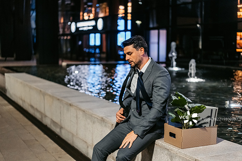 伤心的中年商人坐在公司大楼前，手里拿着一箱办公用品。图片素材