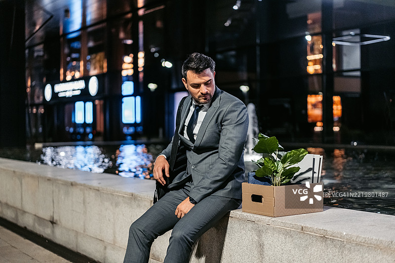 伤心的中年商人坐在公司大楼前，手里拿着一箱办公用品。图片素材