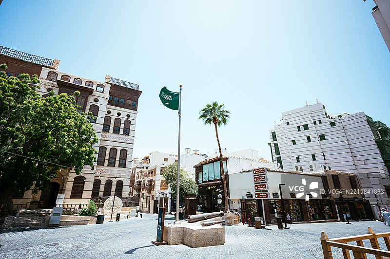 在吉达的历史区阿尔巴拉德，清澈的蓝天下，街景中有沙特阿拉伯国旗、棕榈树和传统建筑。图片素材