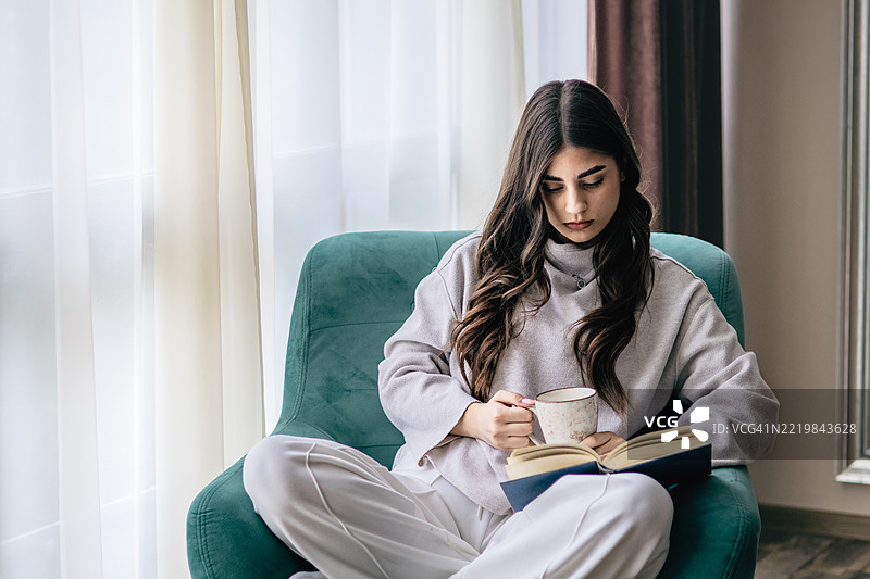 好书、热巧克力和舒适的扶手椅。放松的、迷人的年轻女性，长着棕色的头发，在家中周末阅读书籍。大学生在客厅学习。图片素材