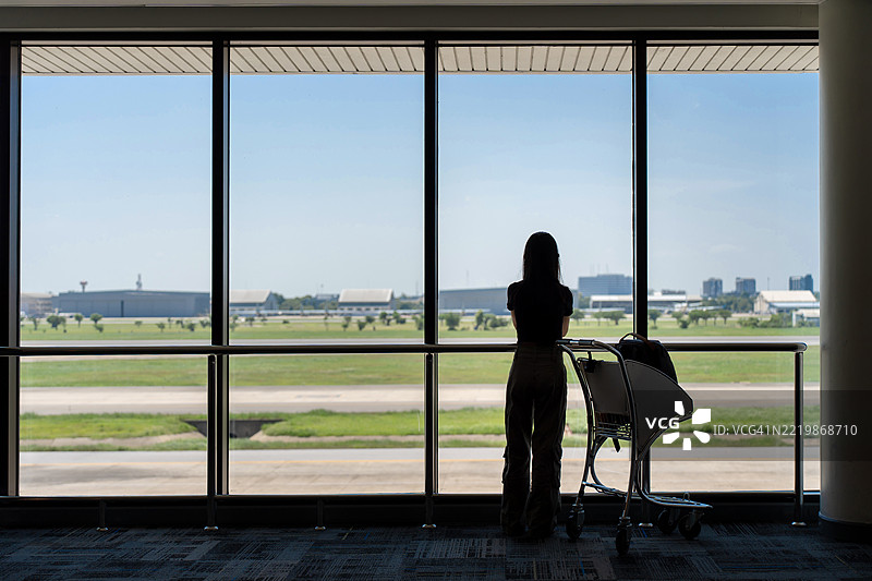亚洲女性在飞机跑道旁观看，身边有行李推车。图片素材