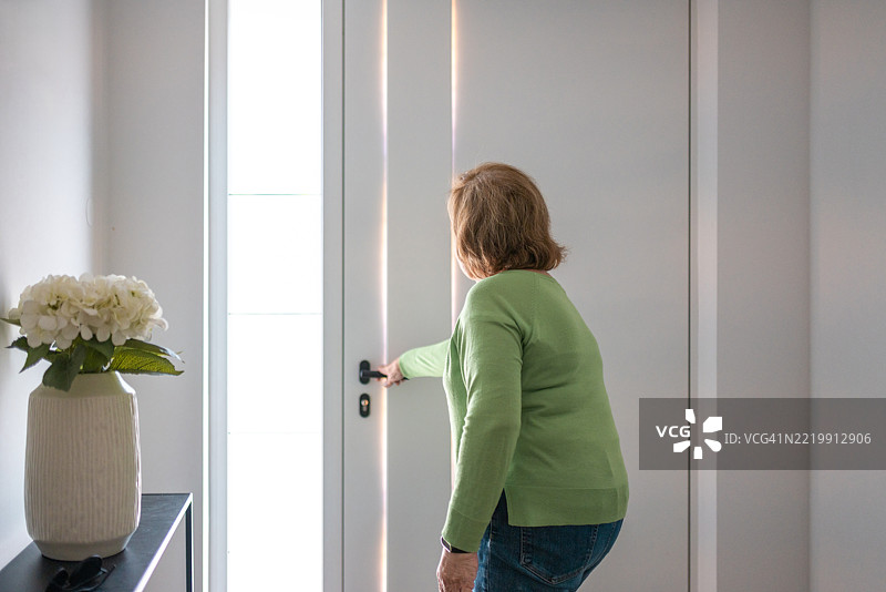 穿着绿色毛衣的老年女性在家中打开一扇明亮的门图片素材