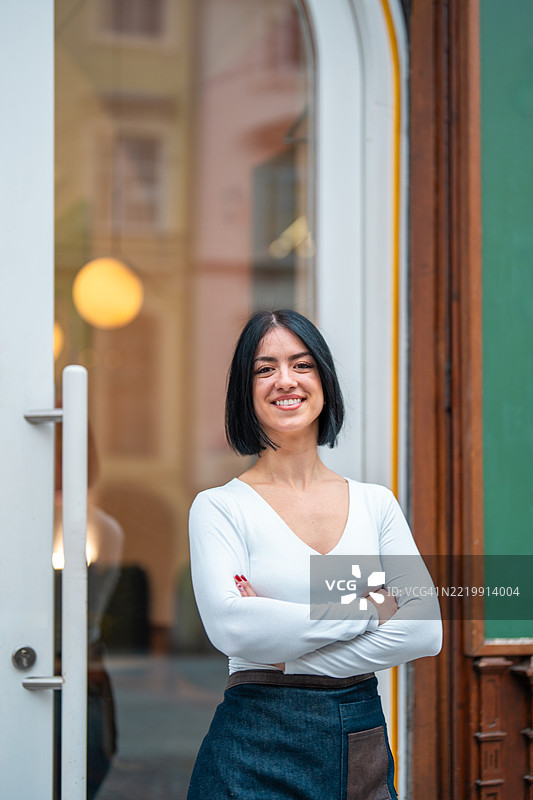 自信的女性咖啡师穿着白色上衣和围裙，站在一家舒适的咖啡馆外。图片素材